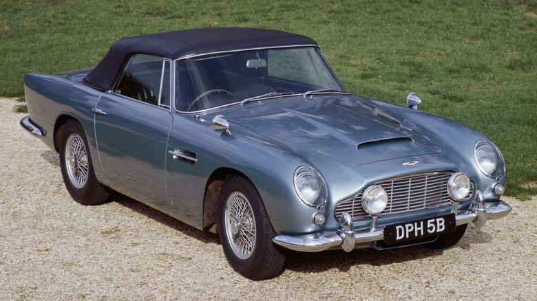 A blue Aston Martin DB5 Convertible is parked on gravel.