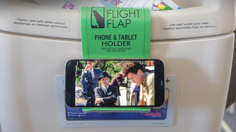 A Flight Flap phone and tablet holder holding a smartphone attached to the back of a passenger seat on an airplane.