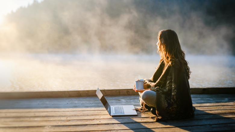 A woman sitting on decking with her laptop looking out over a misty lake.