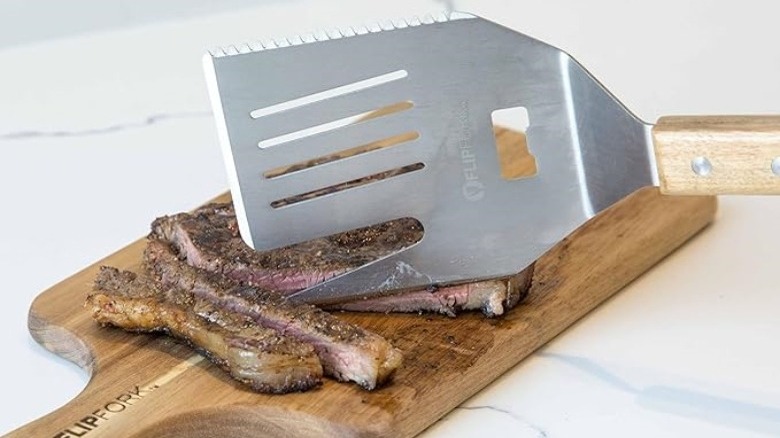 Metal grilling spatula sticking a fork-like edge into a cooked steak on a cutting board