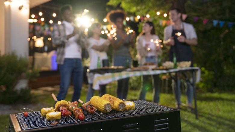 Group of friends in a yard behind a small grill cooktop feature various foods