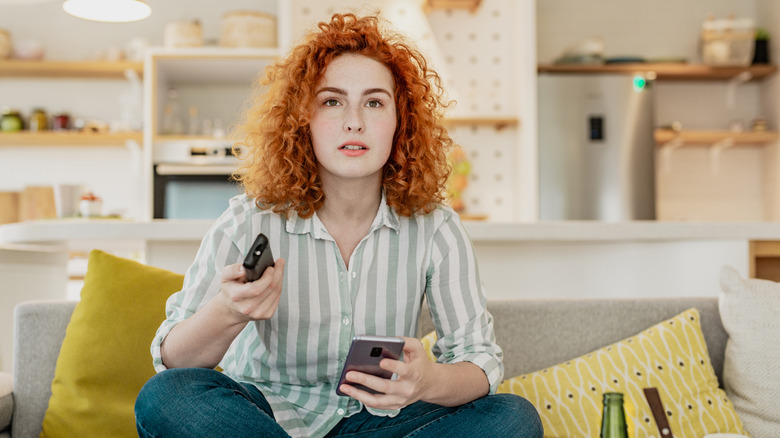 woman holding smartphone and remote control sitting on couch