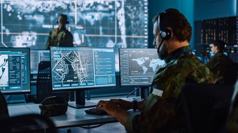 A man in military fatigues sits at a bank of computer monitors