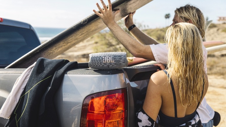 A young man and woman fetch a surfboard from their pickup truck while a JBL Charge 5 rests on the vehicle.