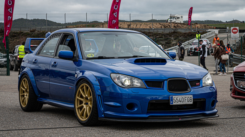 A modified Hawk Eye Subaru Impreza WRX STI at a car show, front 3/4 view, blue exterior with gold wheels