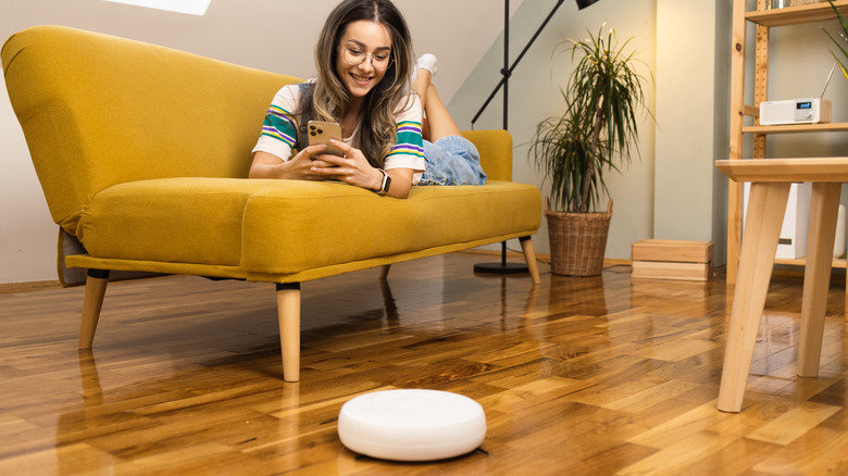 Woman on a yellow sofa holding a smartphone looking at a white smart device on the floor