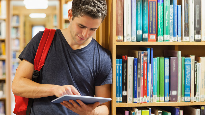 Student holding a tablet pc and leaning against a bookshelf