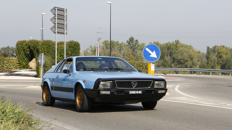 A Lancia Montecarlo in blue with gold wheels driving, front 3/4 view