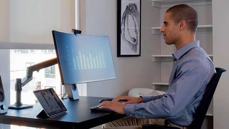 A man sitting at the desk with his large monitor supported by the Ergotron LX Premium Monitor Arm.