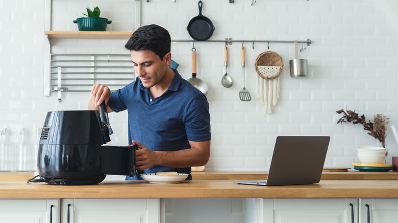 A man uses an air fryer in a kitchen with a laptop open on the counter