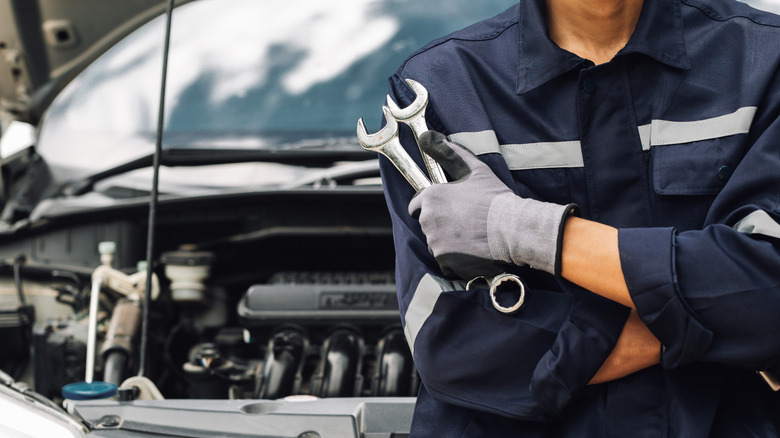 A mechanic holds wrenches near an open car hood.