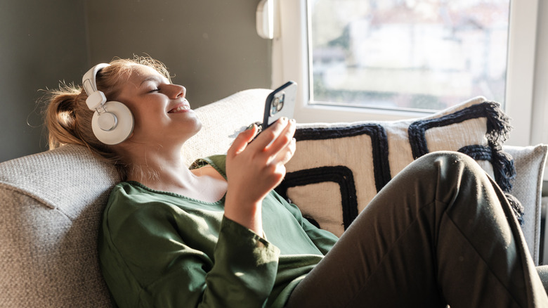 woman on a couch holding a phone and wearing white headphones smiling and listening to music