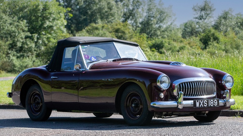 A maroon Nash-Healey car driving on a country road with two people in it.