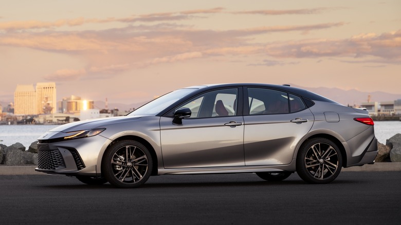 Side view of silver Toyota Camry on a road by a body of water with tall buildings in the background