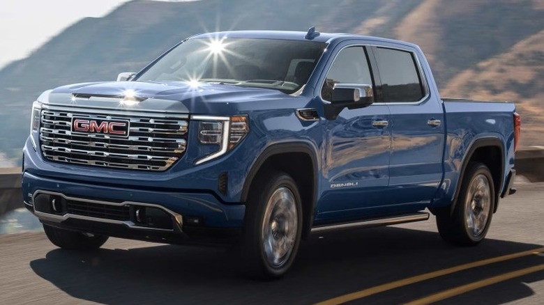 A blue GMC Sierra 1500 Denali driving on a road with mountains in the background.