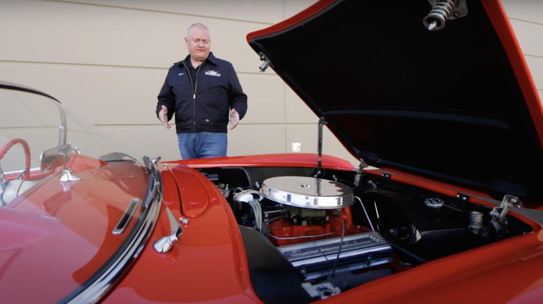 Man in blue jacket demonstrating V8 engine in red 1955 Corvette with hood open