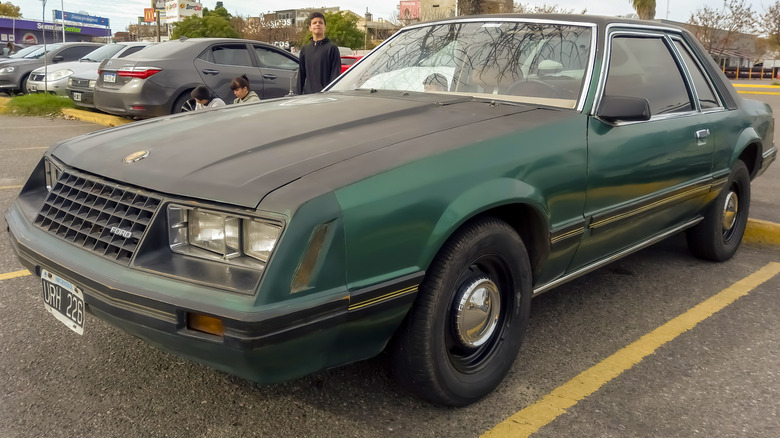 Green 1979 Ford Mustang with black hood in parking lot with. man standing behind