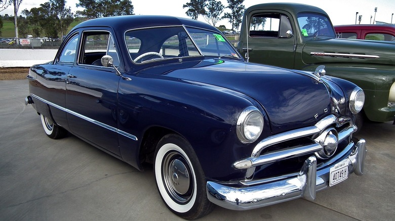 A blue 1949 Ford coupe parked in front of a green vintage pickup truck.