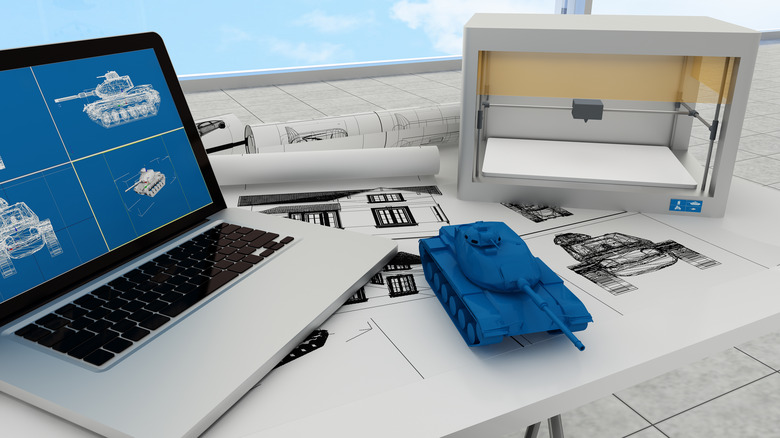 Blueprints of a tank on a laptop screen beside a 3D printer and 3D printed main battle tank.