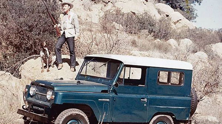 Roy Rogers holding rifle standing on rock behind blue Nissan Patrol