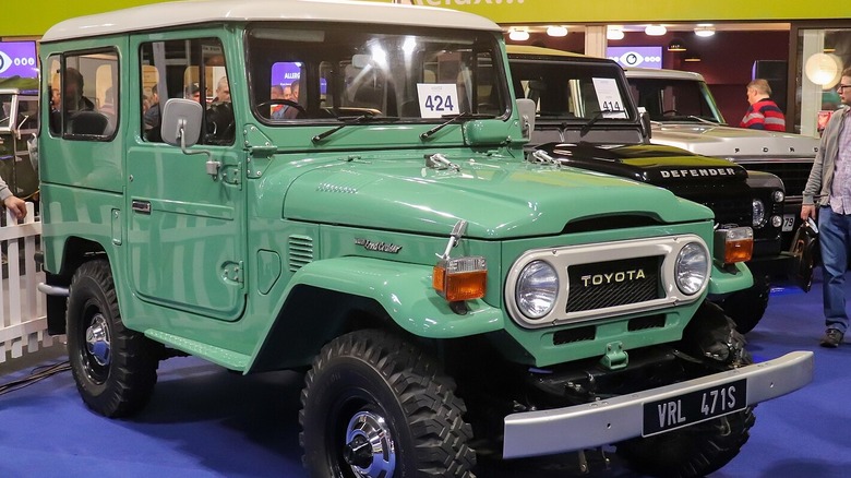 Teal Toyota FJ40 Land Cruiser on blue carpet at car show, Land Rover Defender and Ford Bronco in background