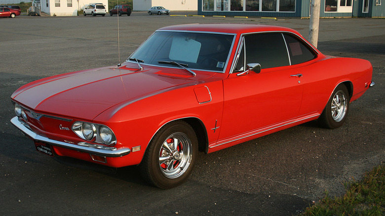 A red Chevrolet Corvair in a nearly empty parking lot with a few other cars in the distance