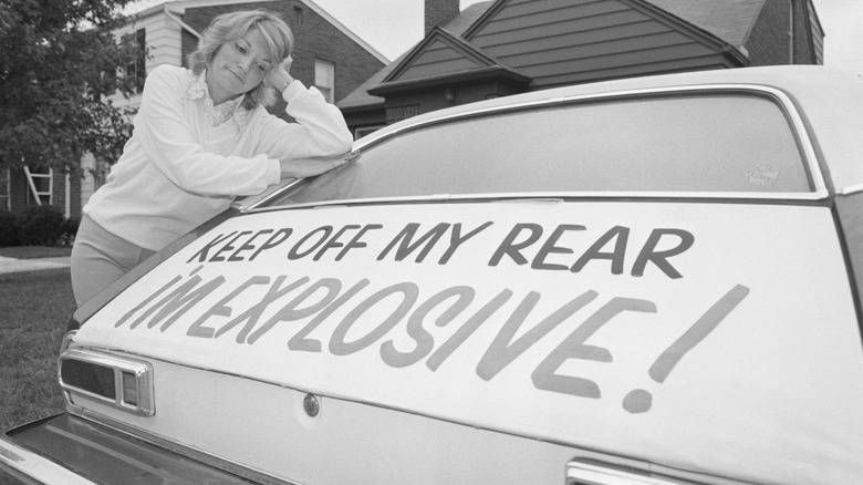 A woman leas on a Ford Pinto that bears a sign reading, "Keep off my rear, I'm explosive!"