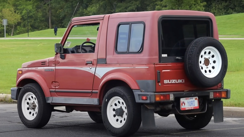 A red Suzuki Samurai hardtop in a parking lot in front of a grassy field