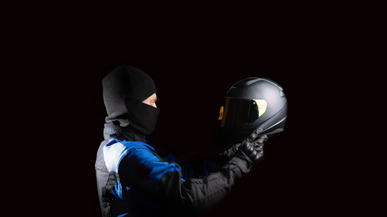 A person holding a helmet while donning a balaclava and a full-body of motorcycle gear