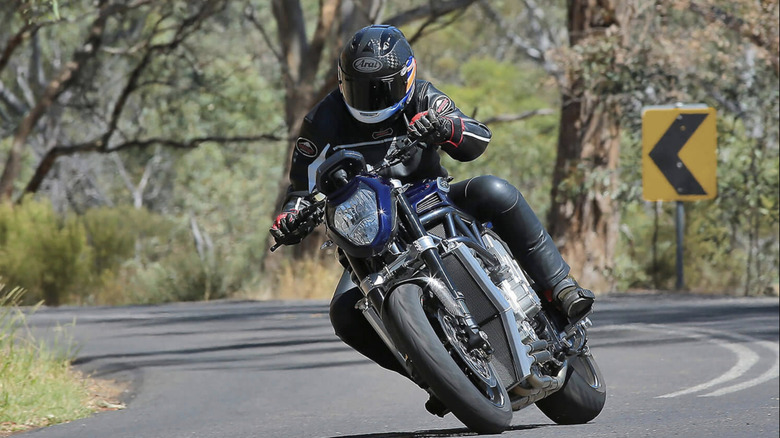 Helmeted rider on PGM V8 motorcycle leaning into right-hand curve on wooded roadway