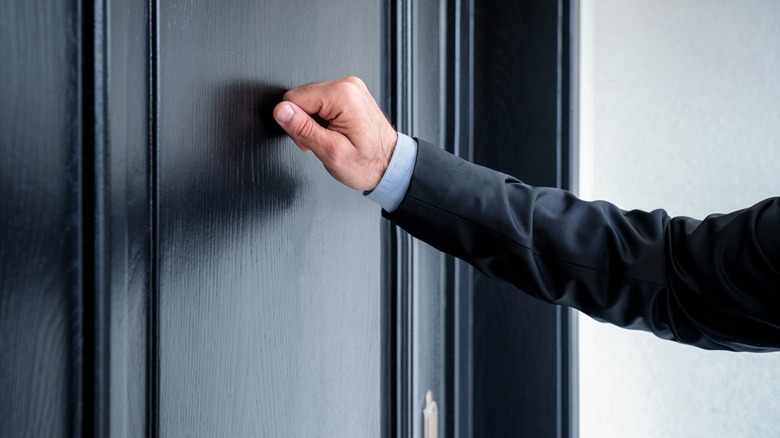 A hand of a man in a charcoal gray suit knocks on a black wooden door