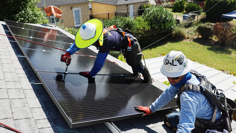 A pair of workers install solar panels on a house's roof with the backyard visible below