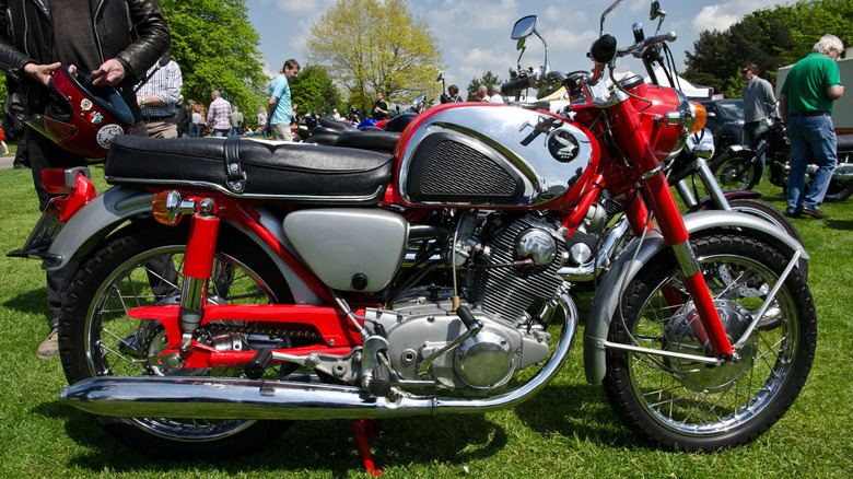 Red and silver Honda CB77 Super Hawk in a field surrounded by people