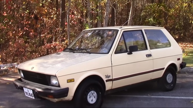 A Light Colored Yugo Parked In Front Of A Forest