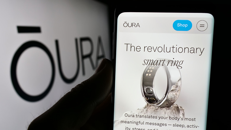 A hand holding a smartphone displaying information about the Oura RIng as the company's logo is flashed on a big screen in front