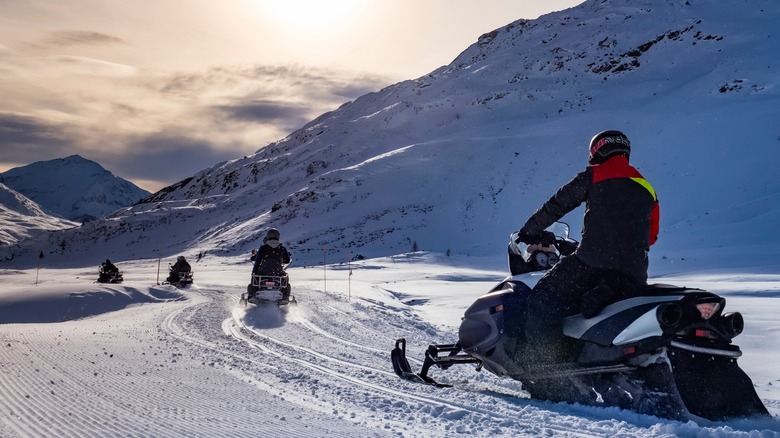 Row of snowmobile riders pass mountain