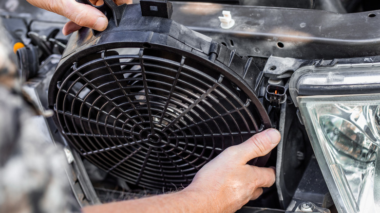 A man inserts an engine cooling fan under the radiator grill of a car.