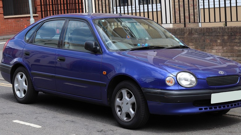 Blue 1998 Toyota Corolla parked on street in front of brick wall and iron fencing