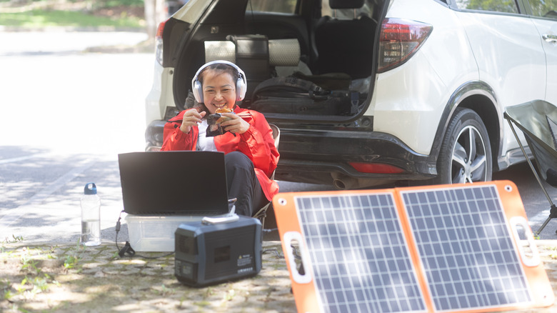 Woman with headphones viewing her laptop while smiling, SUV is in the background while solar panel and battery bank are in the foreground.