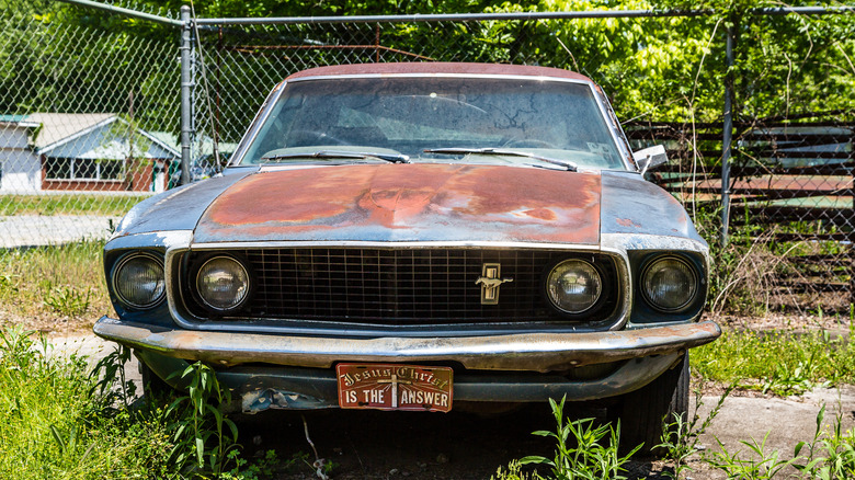 A rusty Ford Mustang in front of a chain-link fence