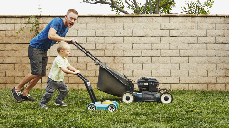 Adult mowing the lawn with a child using a toy mower next to them