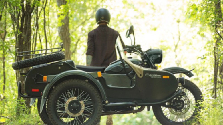 Rider standing by Ural Sportsman in woods looking toward the sun