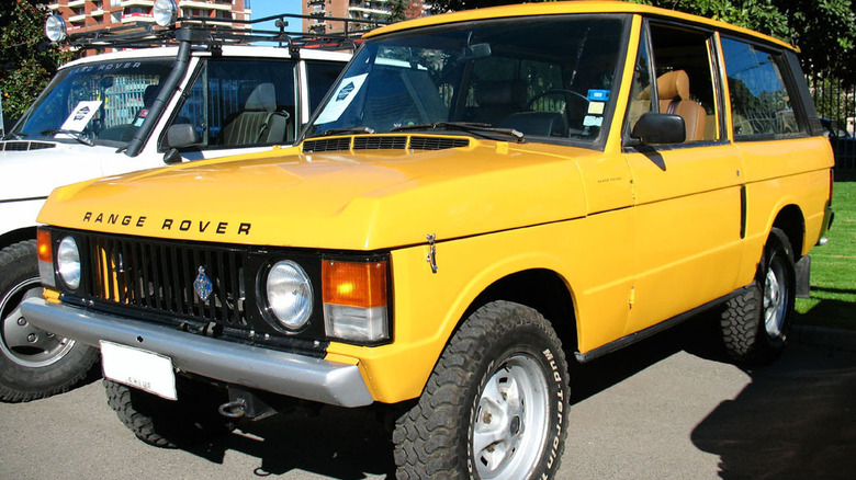 Yellow 1979 Range Rover parked in lot with white Rover and grass in background