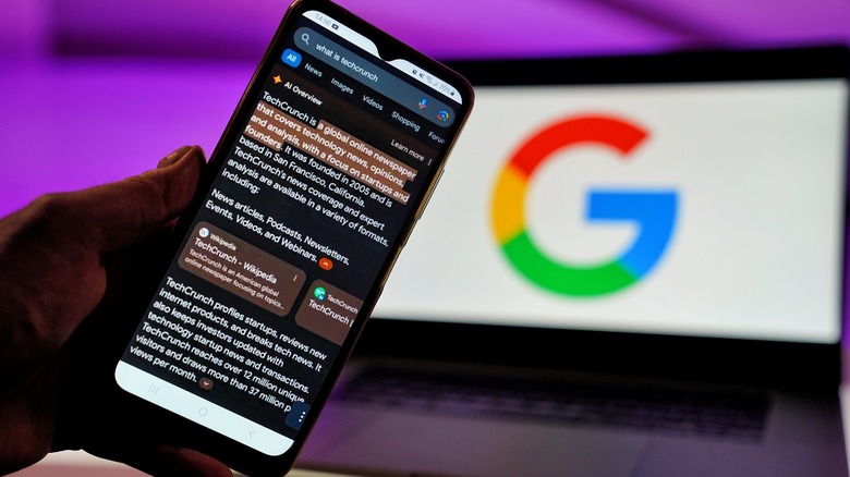 Google AI Overview on a smartphone screen in front of a monitor out of focus showing the Google logo