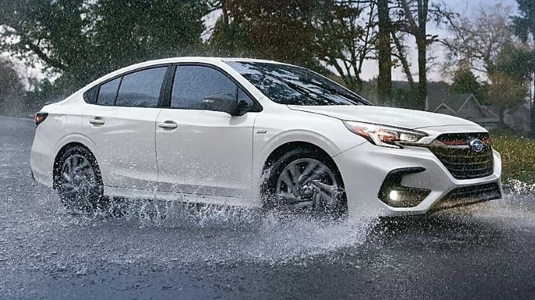 Crystal white pearl Subaru Legacy Sport driving through a large puddle with trees in the background