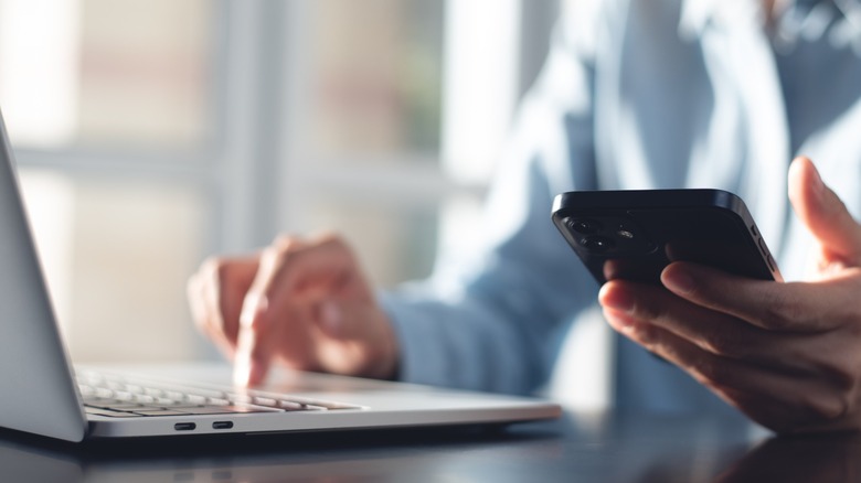 A person holding a phone and touching a laptop