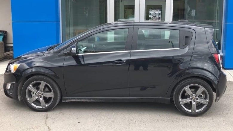 Side profile shot of a black 2015 Chevrolet Sonic RS parked on a paved surface right in front of a building