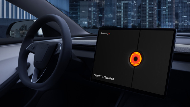 Tesla 3 dashboard showing steering wheel and digital screen. The digital screen displays a logo and the words "Sentry activated". The photo is taken at night with a city scene visibe through the windscreen.