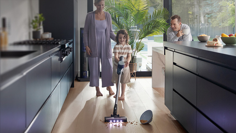 A child uses the Tineco vacuum to clean up spilled pet food in the kitchen as Mom and Dad watch, smiling