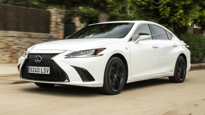 White Lexus ES 300h F Sport driving down a road next to a building with trees